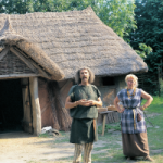 Photograph of a man and a woman dressed as ancient Britons standing in front of a reconstructed British home.