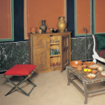 Photography of the interior of room in a Roman house. A table with various bowls on it, a cupboard containing numerous vessels, a couch and a stool are visible against the backdrop of a decorated wall.